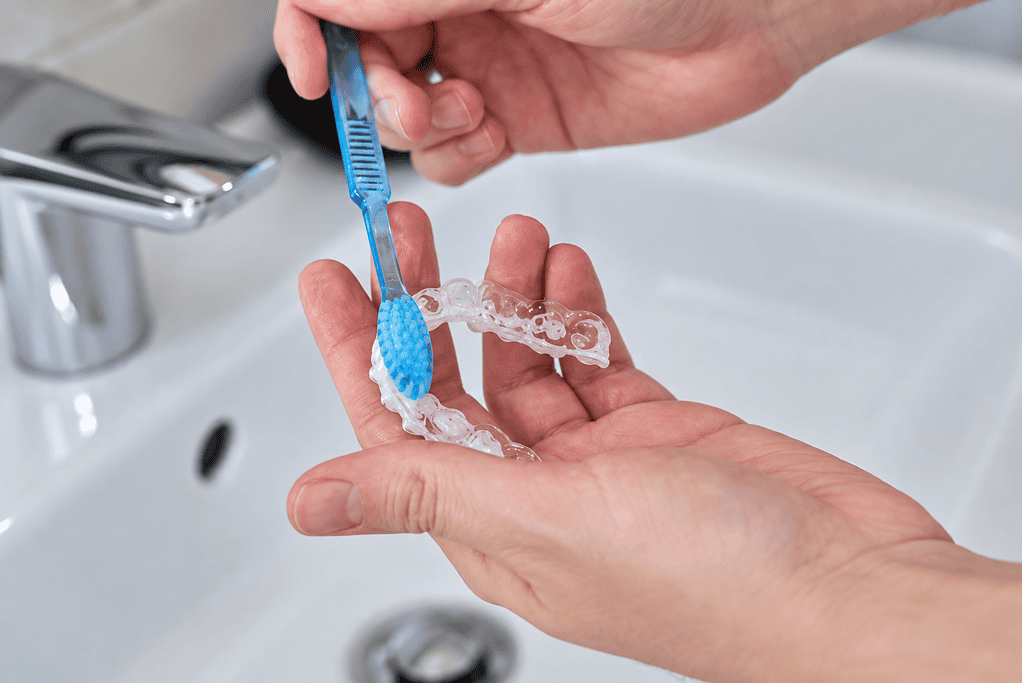 Person cleaning dental aligner with toothbrush under running water.